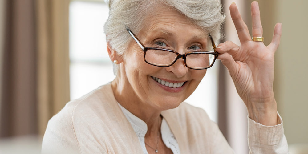 Happy retired senior woman looking at camera while holding eyeglasses. Smiling satisfied woman wearing spectacles at home. Closeup face of old grandmother trying on new eyewear.