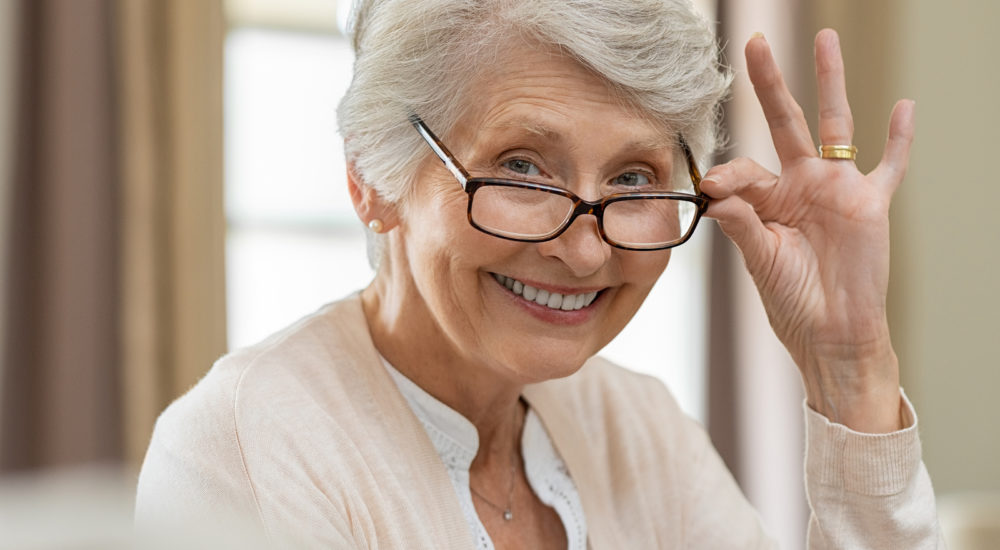 Happy retired senior woman looking at camera while holding eyeglasses. Smiling satisfied woman wearing spectacles at home. Closeup face of old grandmother trying on new eyewear.